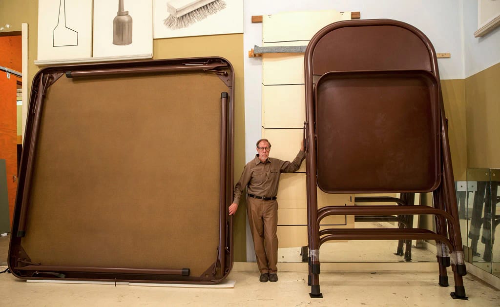 Robert Therrien stojaci pri zväčšenej skladacej súprave stola a stoličiek vo svojom štúdiu v Los Angeles, ukazujúci surrealistickú monumentalitu a funkčnosť objektu.