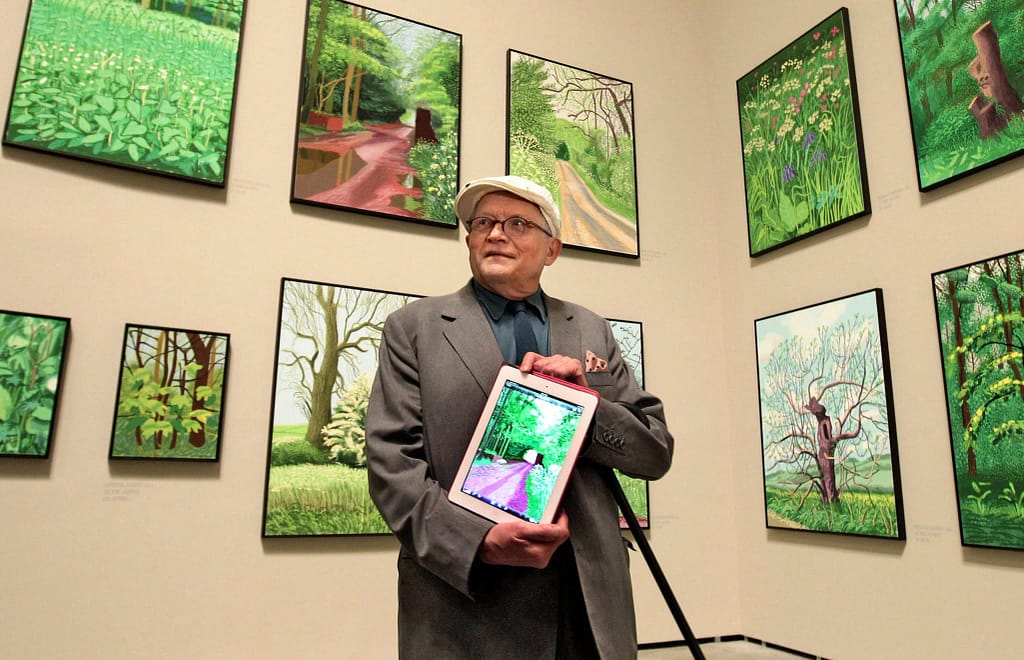 David Hockney drží iPad obklopený farebnými krajinomalbami stromov na výstave v Guggenheim Museum Bilbao, 2012