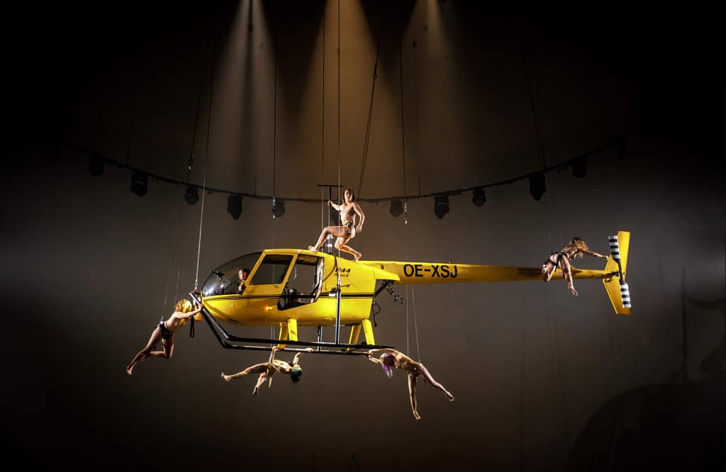 Záber z performancie Florentiny Holzinger Ophelia's Got Talent, 2022 — performerky zavesené na žltom helikoptére nad javiskom Volksbühne v Berlíne.