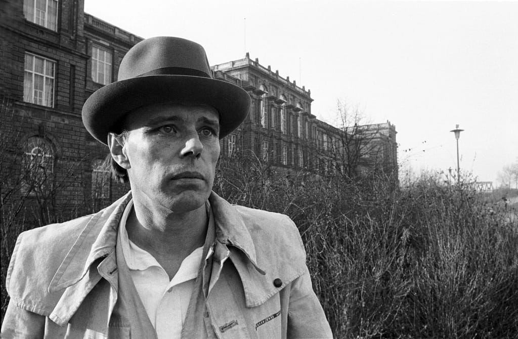 Joseph Beuys v charakteristickom plstenom klobúku a veste, Düsseldorf. V pozadí budova Kunstakademie Düsseldorf a Rheinturm.