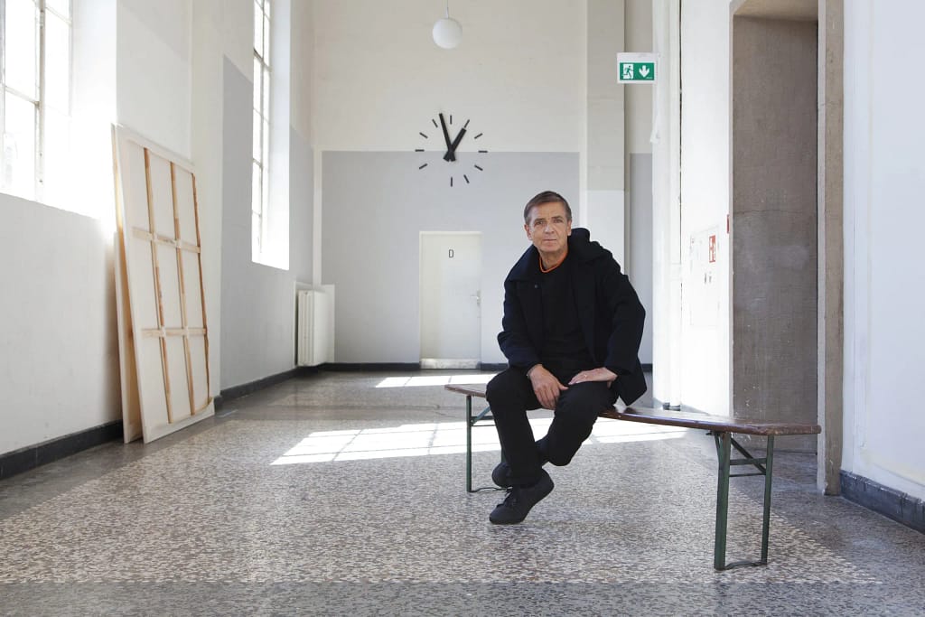 Andreas Gursky sedí v minimalistickom štúdiu na Kunstakademie Düsseldorf, biele steny, prázdne plátna, hodiny na stene