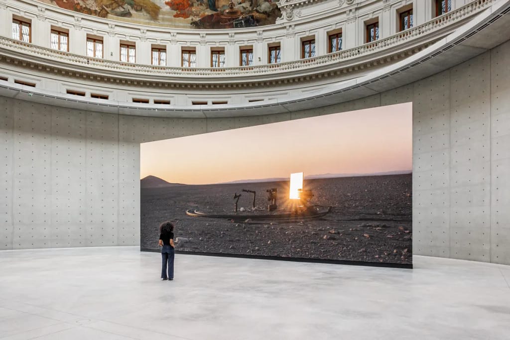 Inštalácia Pierre Huyghe Camata, 2024 — filmová projekcia v rotunde Bourse de Commerce, Paríž, s postavou sledujúcou západ slnka nad krajinou.