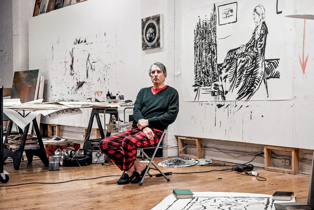 Raymond Pettibon sedí v ateliéri obklopený kresbami a pracovným stolovým materiálom, Manhattan, New York.