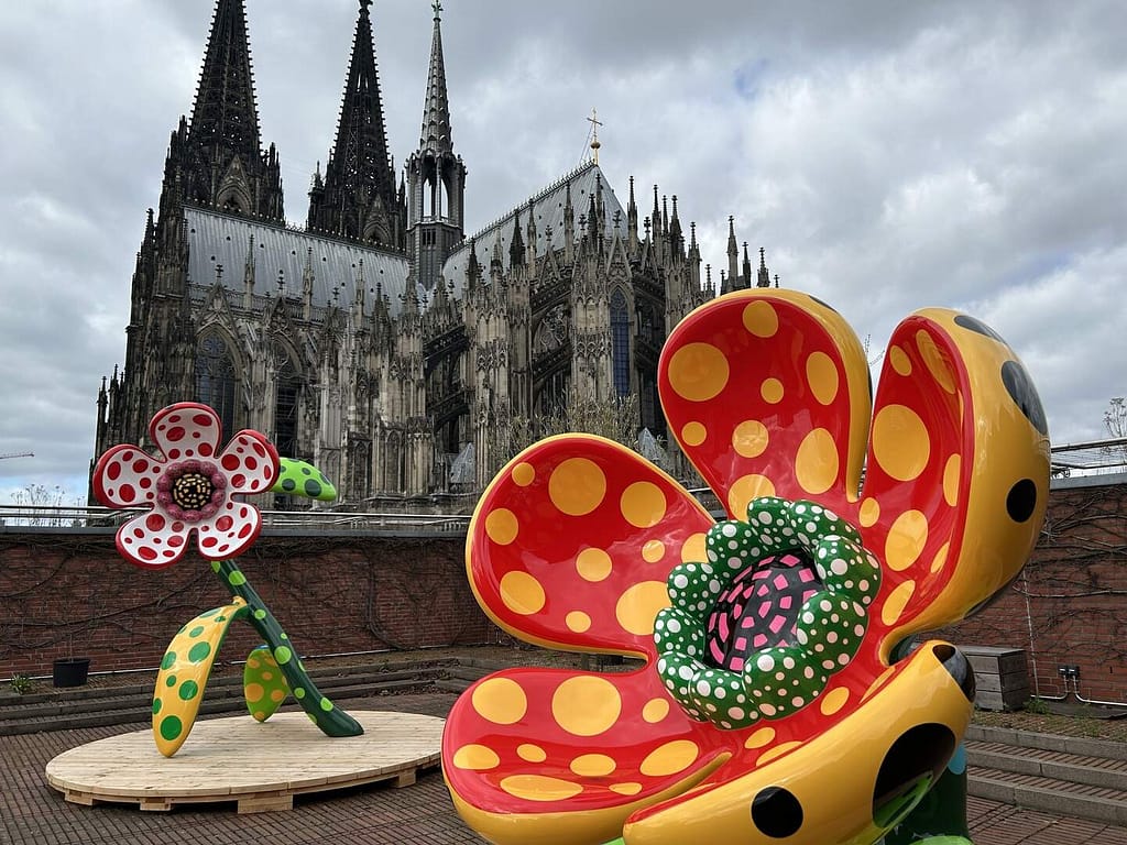 Farebné kvetinové skulptúry Yayoi Kusamy s bodkami na streche Museum Ludwig, v pozadí Kolínsky dóm, 2026