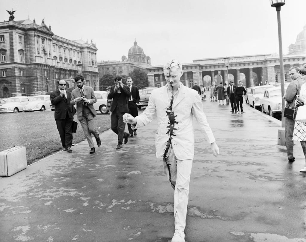 Günter Brus kráča ulicou Viedne v bielom obleku pomaľovanom čiernou čiarou počas akcie Wiener Spaziergang, 1965.