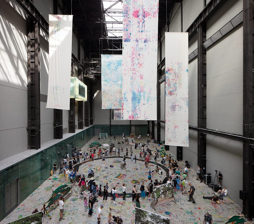 Inštalačný pohľad na dielo Eia Arakawu-Nasha Mega Please Draw Freely v Tate Modern v Londýne, 2021 — veľké maľované plátna visiace zo stropu, návštevníci kreslia na podlahu.